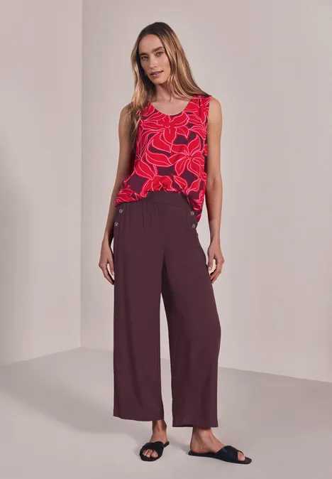 High Waist Wide Leg Hose im Loose Fit rustic rouge Frau trägt rotes ärmelloses Oberteil mit Blütendruck, weite bordeauxrote Hose und schwarze Sandalen.