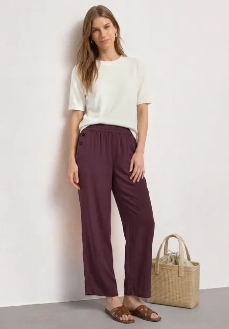 High Waist Wide Leg Hose im Loose Fit rustic rouge Dame trägt weißes T-Shirt und weinrote Hose, Sandalen, neben ihr eine Basttasche.