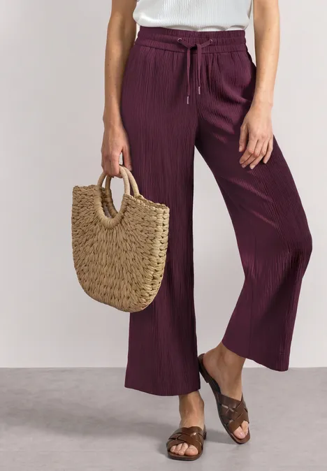Mid Waist Wide Leg Hose im Loose Fit rustic rouge Frau trägt lila weite Hose mit weißem Top, braunen Sandalen und geflochtenen Basttasche.