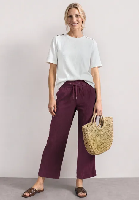 Mid Waist Wide Leg Hose im Loose Fit rustic rouge Person trägt weißes T-Shirt, weite bordeauxrote Hosen, schwarze Sandalen und hält eine geflochtene Korbtasche.