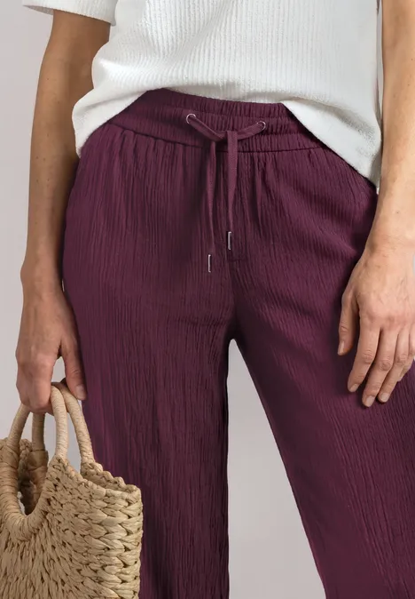 Mid Waist Wide Leg Hose im Loose Fit rustic rouge Weite, gerippte bordeauxrote Stoffhose mit Tunnelzug, weißer Top und Korbtasche.