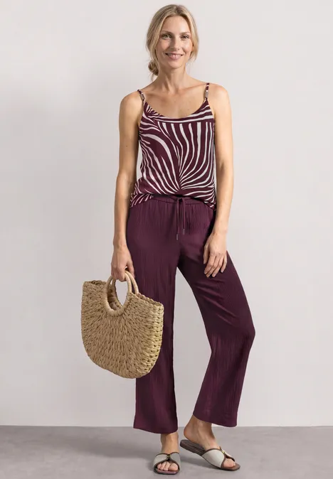Mid Waist Wide Leg Hose im Loose Fit rustic rouge Frau trägt gestreiftes Top, breite lila Hose, Sandalen und eine Strohtasche.