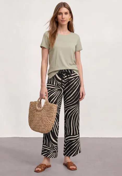 7/8 Wide Leg Hose im Loose Fit Black Frau mit grüner Kurzarmbluse, schwarz-weißen Zebramuster-Hosen, Sandalen und Strohtasche vor neutralem Hintergrund.