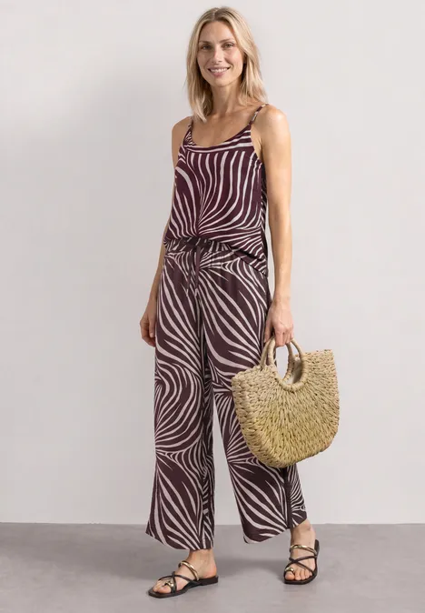 Vrouw in zebraprint top en broek met een strooien tas en zwarte sandalen, tegen een neutrale achtergrond.