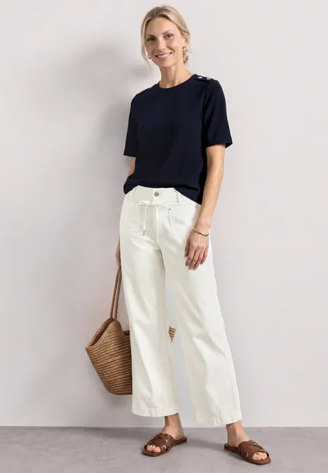 Straight Leg Hose in Washed-Optik off white Person in schlichtem dunkelblauen T-Shirt, weiter weißer Hose, braunen Sandalen und geflochtener Strohtasche.
