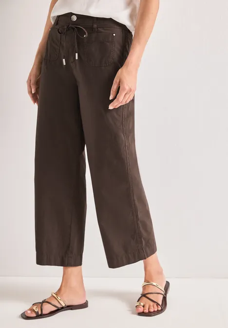 Weite, knöchellange, dunkelbraune Stoffhose mit Bindegürtel, kombiniert mit flachen Riemchensandalen.