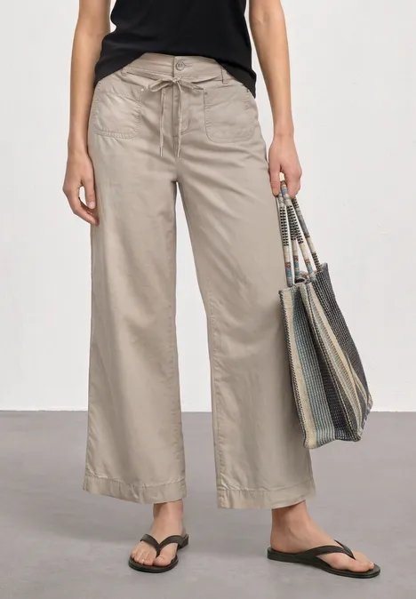 Straight Leg Hose in Washed-Optik sepia sand Frau trägt weite hellbeige Hose, schwarzes Top, Flip-Flops und hält eine gestreifte Tragetasche.