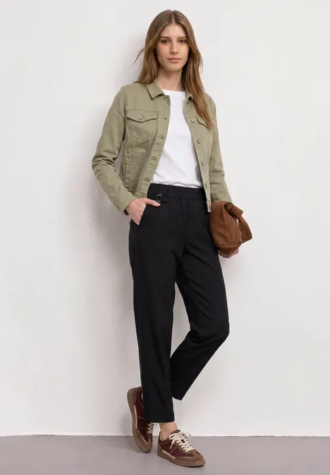 Persoon voor witte muur met groene jas, wit shirt, zwarte broek, bruine schoenen en tas.