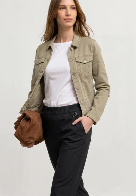 Vrouw in lichtbeige jack, wit T-shirt en zwarte broek, houdt een bruine tas, hand in de zak.