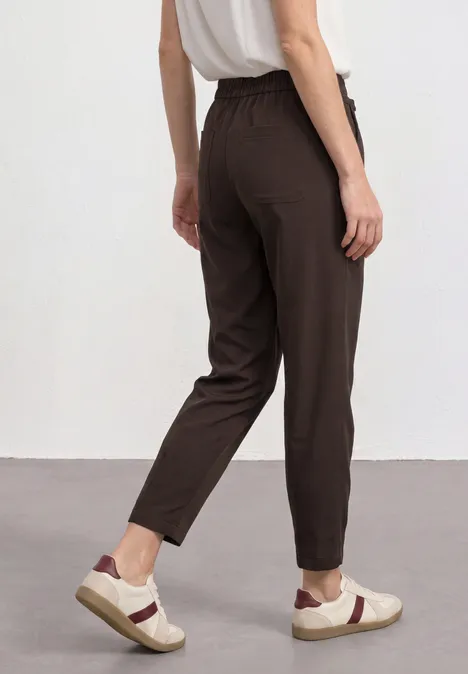 Vrouw van achteren in een losvallende bruine broek, wit hemdje en witte sneakers voor een neutrale achtergrond.