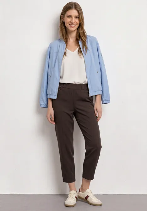 Volledig portret van een vrouw in witte blouse, lichtblauw jasje, bruine chino's en lichte loafers voor een witte muur