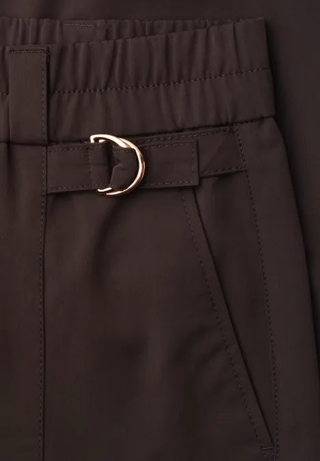 Close-up van een donkerbruine broek met elastische taille, steekzak en goudkleurige D-ring gesp.