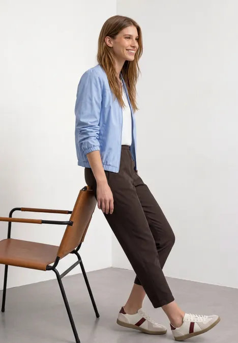 Vrouw in lichtblauw shirt, wit T-shirt, bruine broek en sneakers zit zijdelings op stoel voor witte achtergrond.