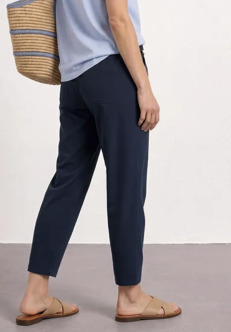 Vrouw draagt donkerblauwe broek, lichtblauw topje, strooien tas en platte sandalen.