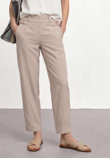 Benen van een vrouw in beige broek, wit topje, sandalen en nonchalante geruite tas.