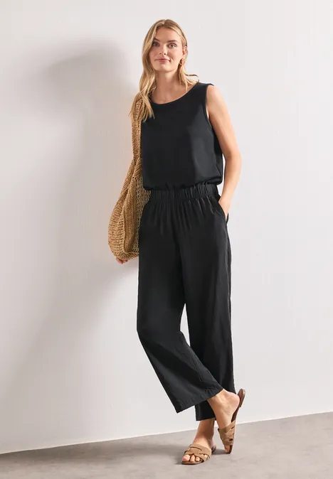 Vrouw in loszittende zwarte jumpsuit met zakken, strooien tas en sandalen voor een witte muur.
