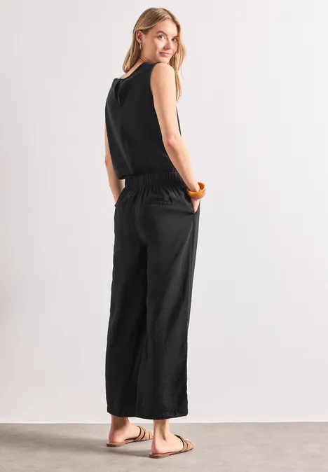 Vrouw in een zwarte mouwloze jumpsuit met wijde pijpen en sandalen, zijaanzicht op neutrale achtergrond.