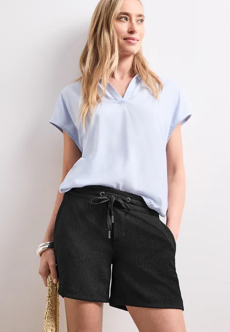 Korte broek met structuur in Loose Fit Black Vrouw in lichtblauw bloesje, zwarte short en met strooien tas.