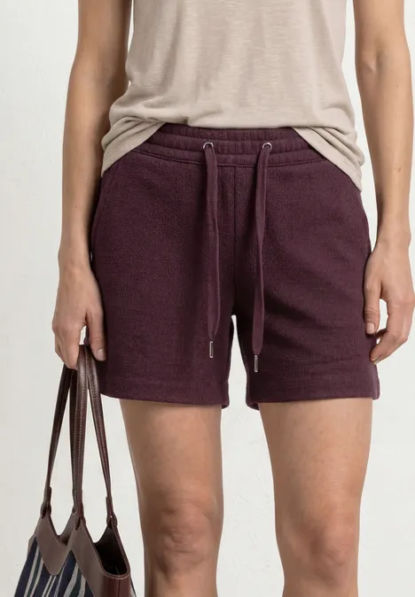 Korte broek met structuur in Loose Fit rustic rouge Persoon draagt een beige top en donkerpaarse sweatshorts met trekkoord, houdt een bruine tas vast.