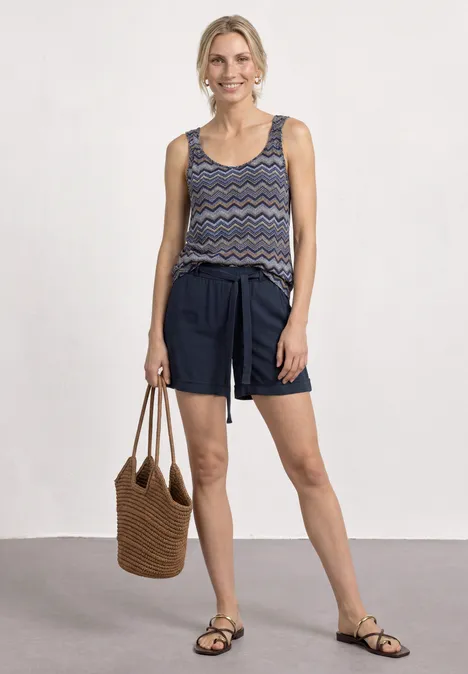 Vrouw met dessin top, donkerblauwe shorts, sandalen en gevlochten tas.