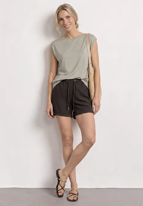 Frau vor weißer Wand, trägt graues Top, schwarze Shorts, Sandalen und Tasche.