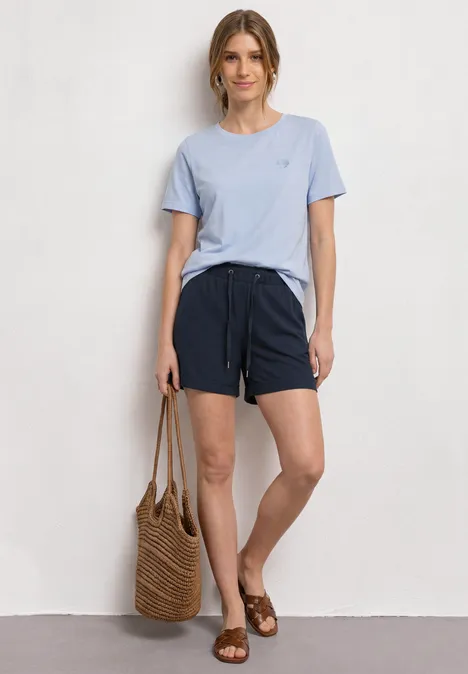 Frau lehnt an Wand, trägt hellblaues Shirt, marineblaue Shorts und Sandalen, hält Strohtasche.