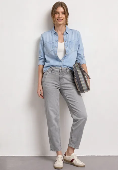 High Waist Barrel Leg Jeans im Loose Fit Light Grey Random Frau vor heller Wand, trägt hellblaues Hemd über weißem Top, graue Hose und helle Schuhe, hält eine Tasche.
