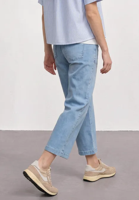 Personne en jean bleu clair et baskets beiges, debout de façon décontractée devant un fond neutre.