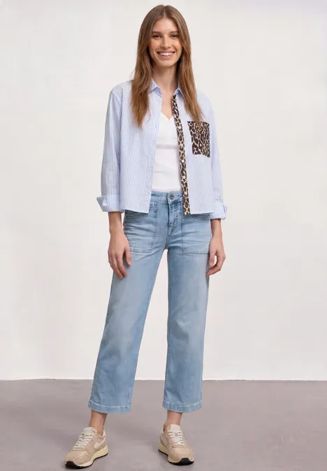 Femme en chemise bleu clair à poche imprimée, top blanc, jean droit bleu et baskets.