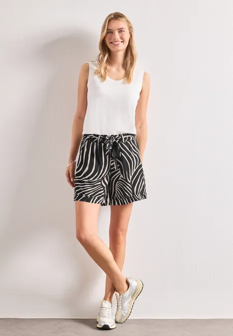 Une jeune femme pose devant un mur blanc avec un débardeur blanc, un short zébré et des baskets.