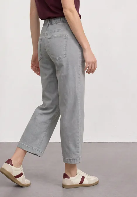 Profil d'une femme en jean gris clair taille haute, coupe large 7/8 et baskets blanches.