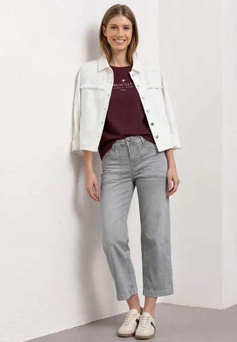 Femme devant un mur blanc, portant une veste blanche, un t-shirt bordeaux, un jean gris droit et des baskets blanches.