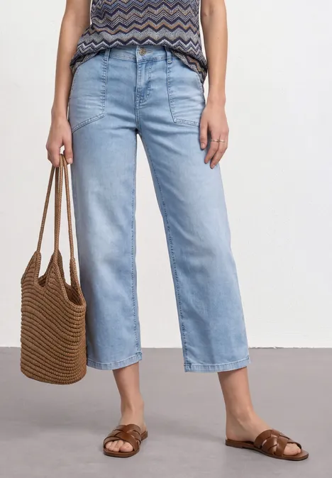 Person trägt hellblaue, gerade geschnittene Jeans, gemustertes Top, Basttasche und geflochtene Sandalen.