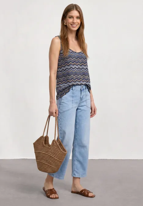 Frau trägt gemustertes ärmelloses Top, weite helle Jeans, braune Sandalen und geflochtene Strohtasche.