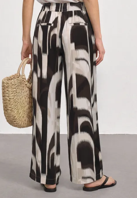 High Waist Wide Leg Hose im Loose Fit espresso brown Frau von hinten in weit geschnittener Hose mit schwarz-weißem abstraktem Muster, trägt Basttasche und flache Sandalen.