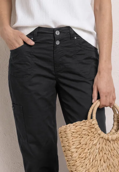 Person trägt ein weißes Top, schwarze Hose mit Knöpfen und hält eine geflochtene Strohtasche.