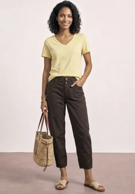 High Waist Barrel Leg Hose espresso brown Frau in gelbem T-Shirt, braunen 7/8-Hosen und Sandalen trägt Korbtasche vor hellem Hintergrund.