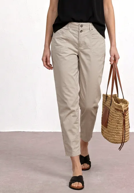 High Waist Barrel Leg Hose clay sand Frau trägt beige, hochgeschnittene Hose, schwarzes Top, Sandalen und eine geflochtene Korbtasche.
