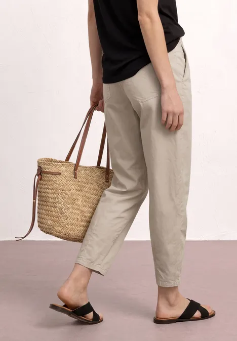 High Waist Barrel Leg Hose clay sand Person trägt beige Hose, schwarze Sandalen und hält einen geflochtenen Strohkörben.