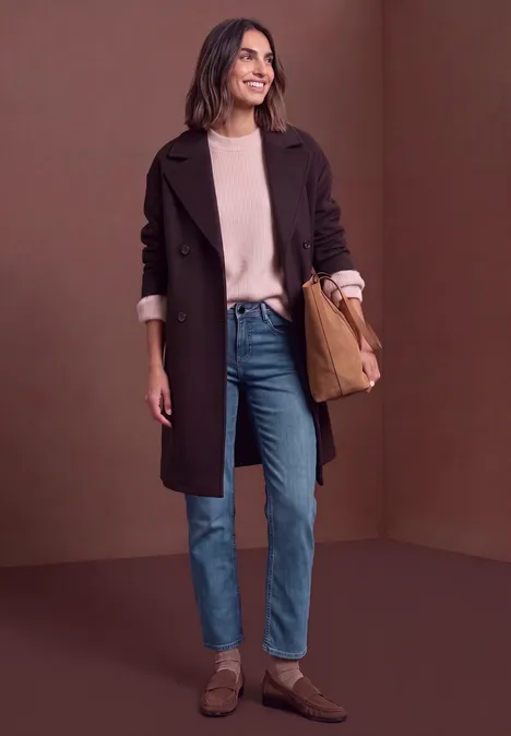 Eine Frau in Mantel, Pullover, Jeans und braunen Schuhen steht vor braunem Hintergrund und lächelt.