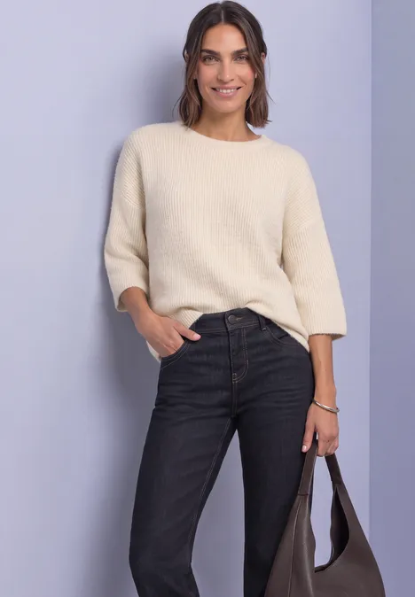 Frau im cremefarbenen Pullover mit 3/4-Ärmeln, schwarzen Jeans und Tasche vor neutralem Hintergrund.