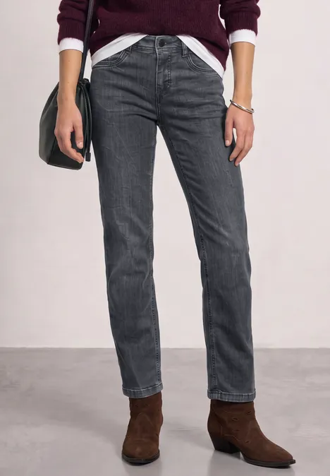 Een persoon draagt donkergrijze jeans en bruine enkellaarsjes bij een wit shirt en een bordeauxrode cardigan.