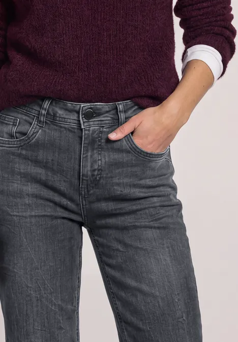 Gedeeltelijke weergave van een persoon in grijze jeans, paarse trui met wit overhemd, hand in zak.