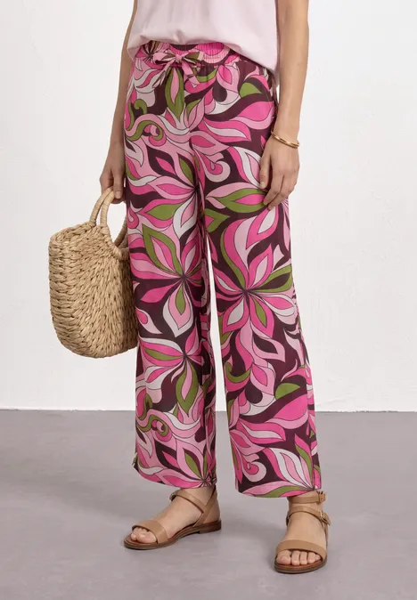 Vrouw draagt ruime roze broek met abstract bladprint, afgewerkt met strozak en sandalen.