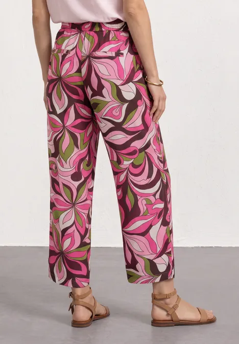 Persoon draagt een broek met sierlijke roze-groene print en platte sandalen voor een witte achtergrond.