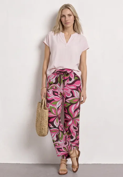 Vrouw in licht t-shirt en losse roze-groene broek met stoffen riem, strooien tas voor witte muur.