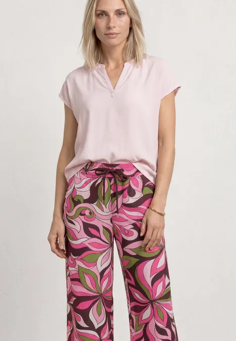 Vrouw draagt lichtroze blouse en losse broek met roze-groene print, staand voor een neutrale achtergrond
