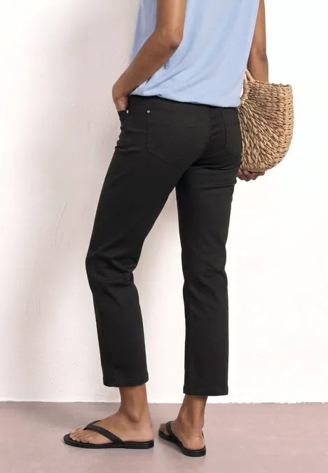 Person von hinten in schwarzer 7/8-Hose, blauem Shirt, mit Strohtasche und schwarzen Sandalen.