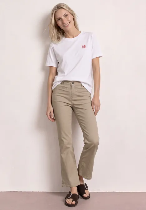Frau lehnt in lässiger Pose an heller Wand, trägt weißes T-Shirt, beigen weiten 7/8-Hose und schwarze Sandalen.