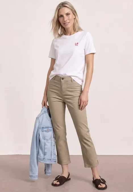 Eine Frau trägt ein weißes T-Shirt mit rotem Herz, beigefarbene Hose, schwarze Sandalen und hält eine Jeansjacke.